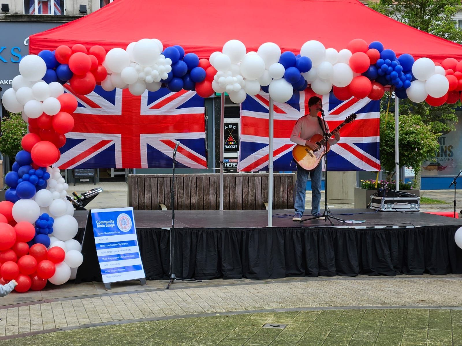 A modular stage with red canopy and red white and blue balloons around it. Solo performer on stage with a guitar