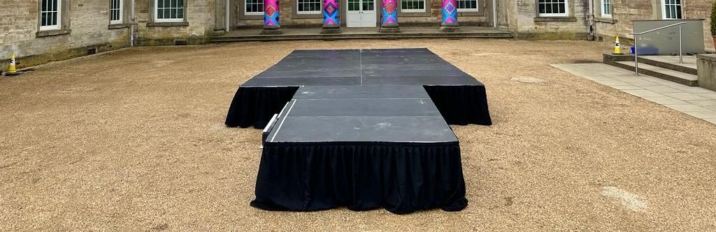 A modular stage set up in a gravel courtyard, with a black skirt
