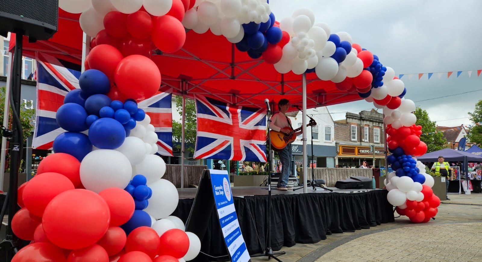 a 6 x 3 metre modular stage with a red canopy and red white and blue balloons arched around the front. A solo performer with his guitar is playing