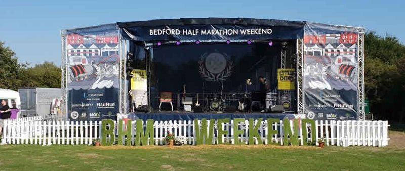 A trailer stage with PA wings set up for the Bedford Half Marathon Weekend. Its branded and there's a white picket fence around the stage