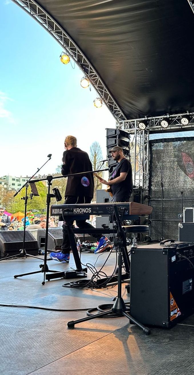Performers on an arc truss stage looking out over an audience with a clear blue sky above them. Production equipment and a piano can be see on stage too. 