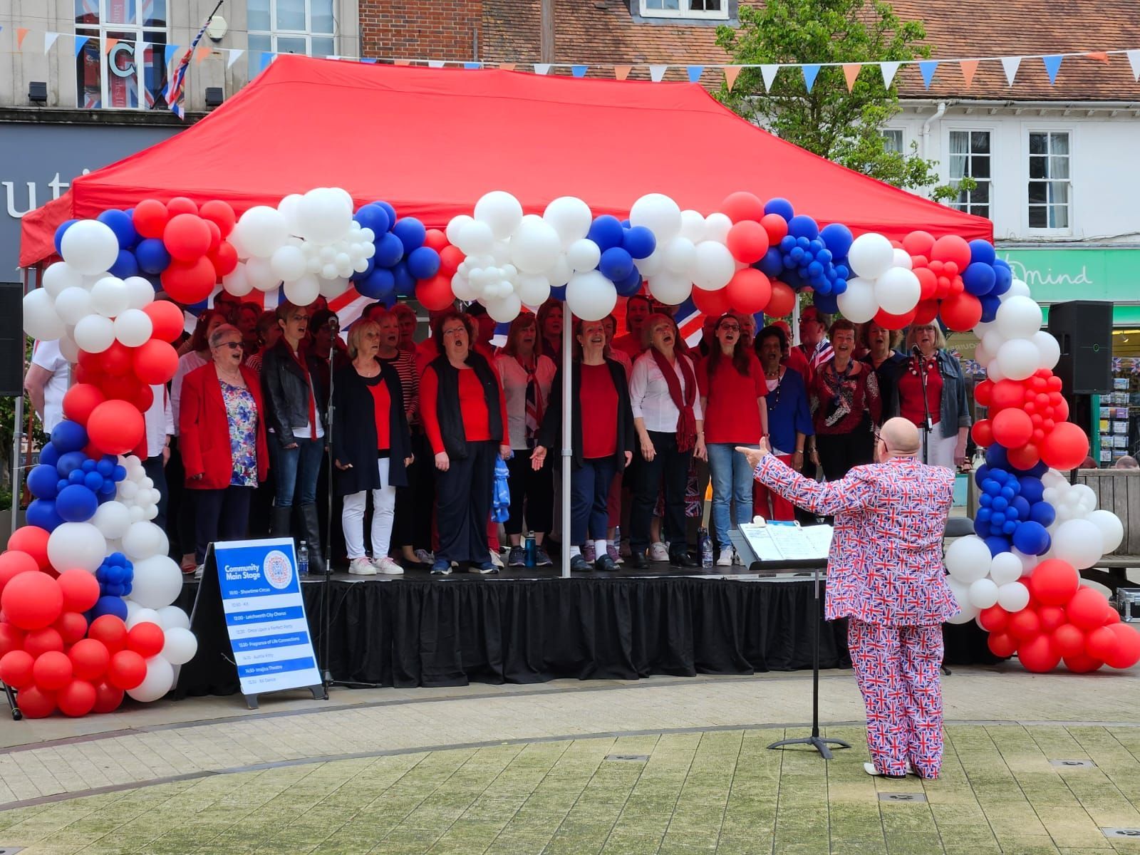 a modular stage with red canopy and red white and blue balloons around it with a choir performing and a conductor wearing pink in front of them