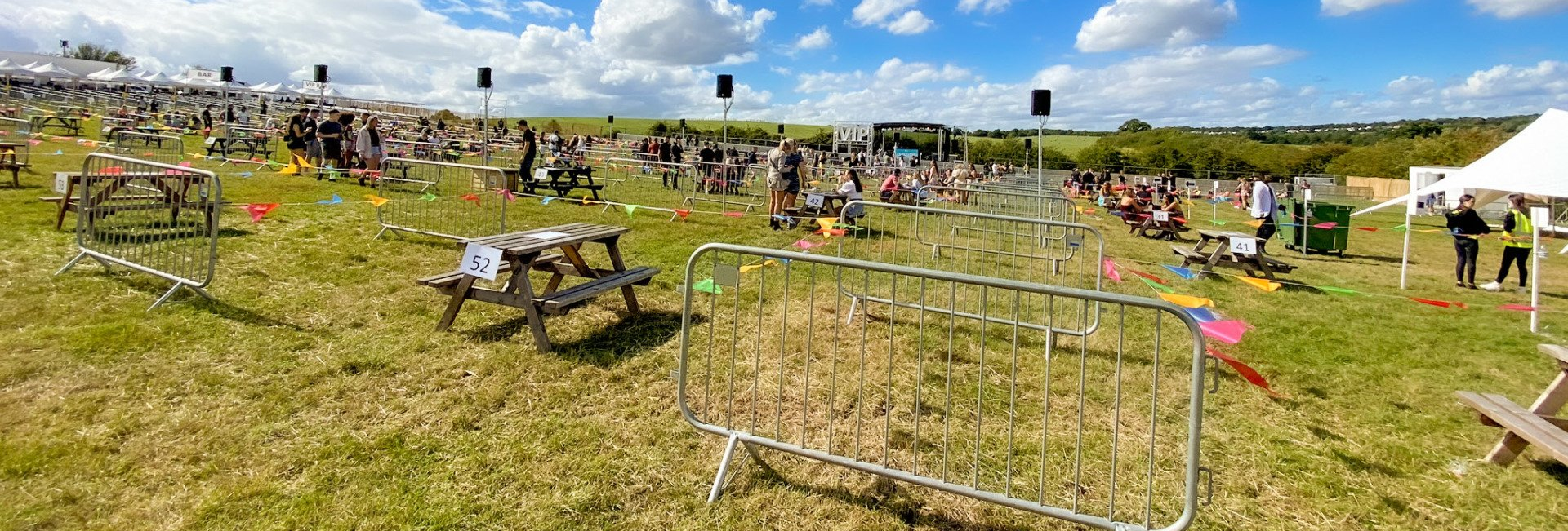 A wide shot of a field set up with picnic tables that are socially distanced. A stage in the background and speakers spread throughout the site.