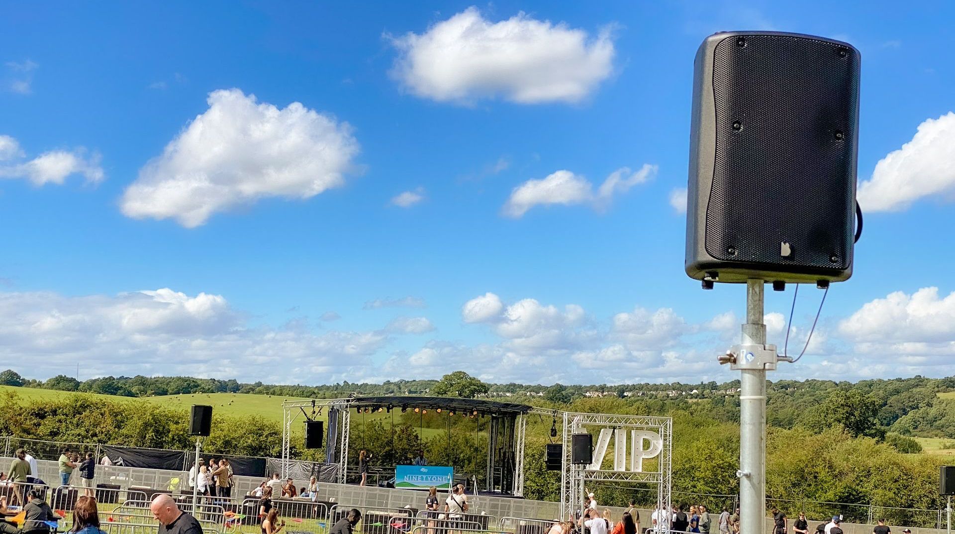 A close up of a speaker on a pole with a trailer stage with PA wings in the background. Picnic tables full of people in front od it socially distanced and a VIP sign.