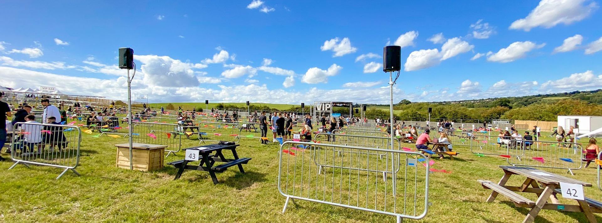 A wide angle of an event site, set up with picnic tables that are socially distances. The trailer stage can be seen in the distance and the sound system throughout the site set up on poles.