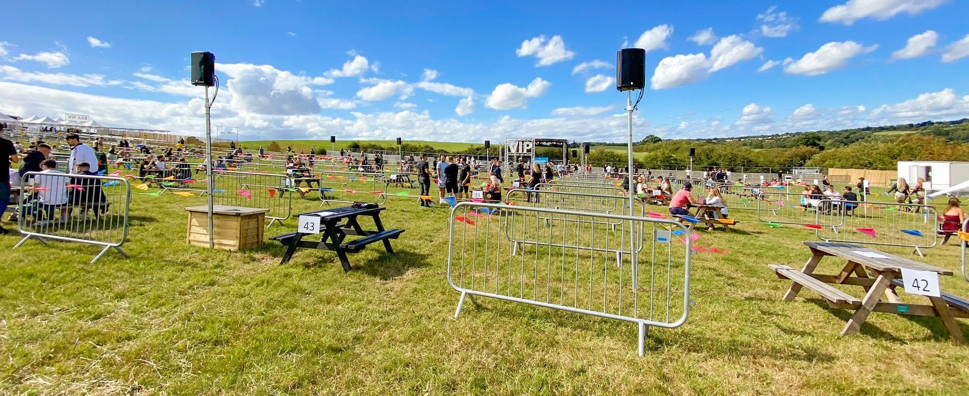 A wide shot of a field set up with picnic tables that are socially distanced. A stage in the background and speakers spread throughout the site.