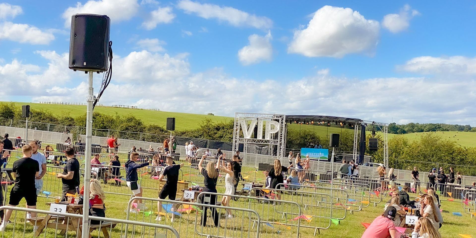 Wide shot of a socially distanced event, with a trailer stage in the background and an audience sat on picnic tables 3 metres apart.