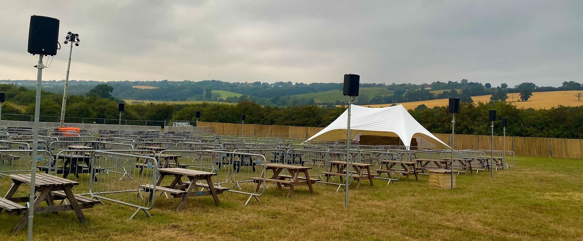 A white stretch tent ina field with picnic tables and crowd barriers set out in front of it socially distanced with speakers on poles around them.