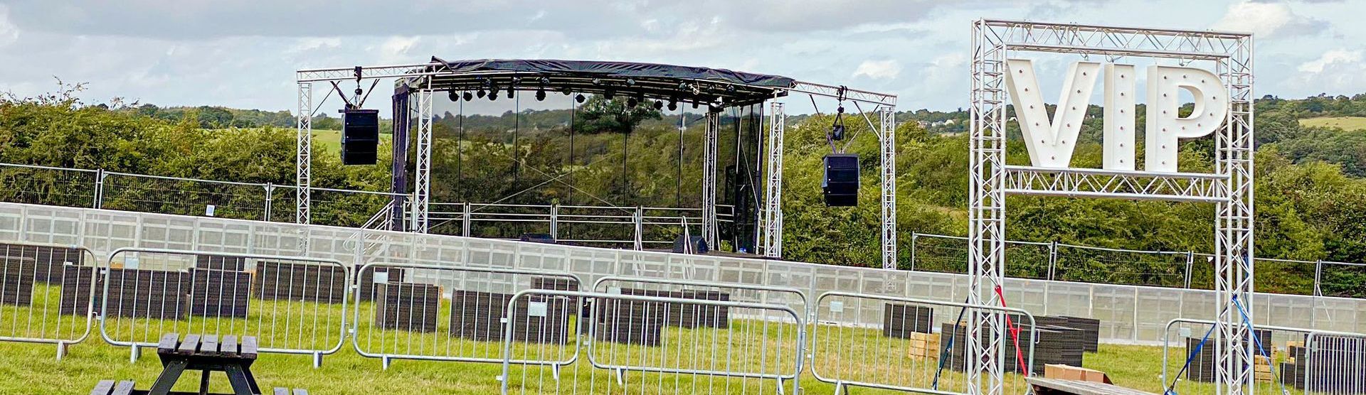 8m x 6m trailer stage with PA wings and a sound rig attached in a field with a view over forests, with a large VIP sign and crowd barriers around it.