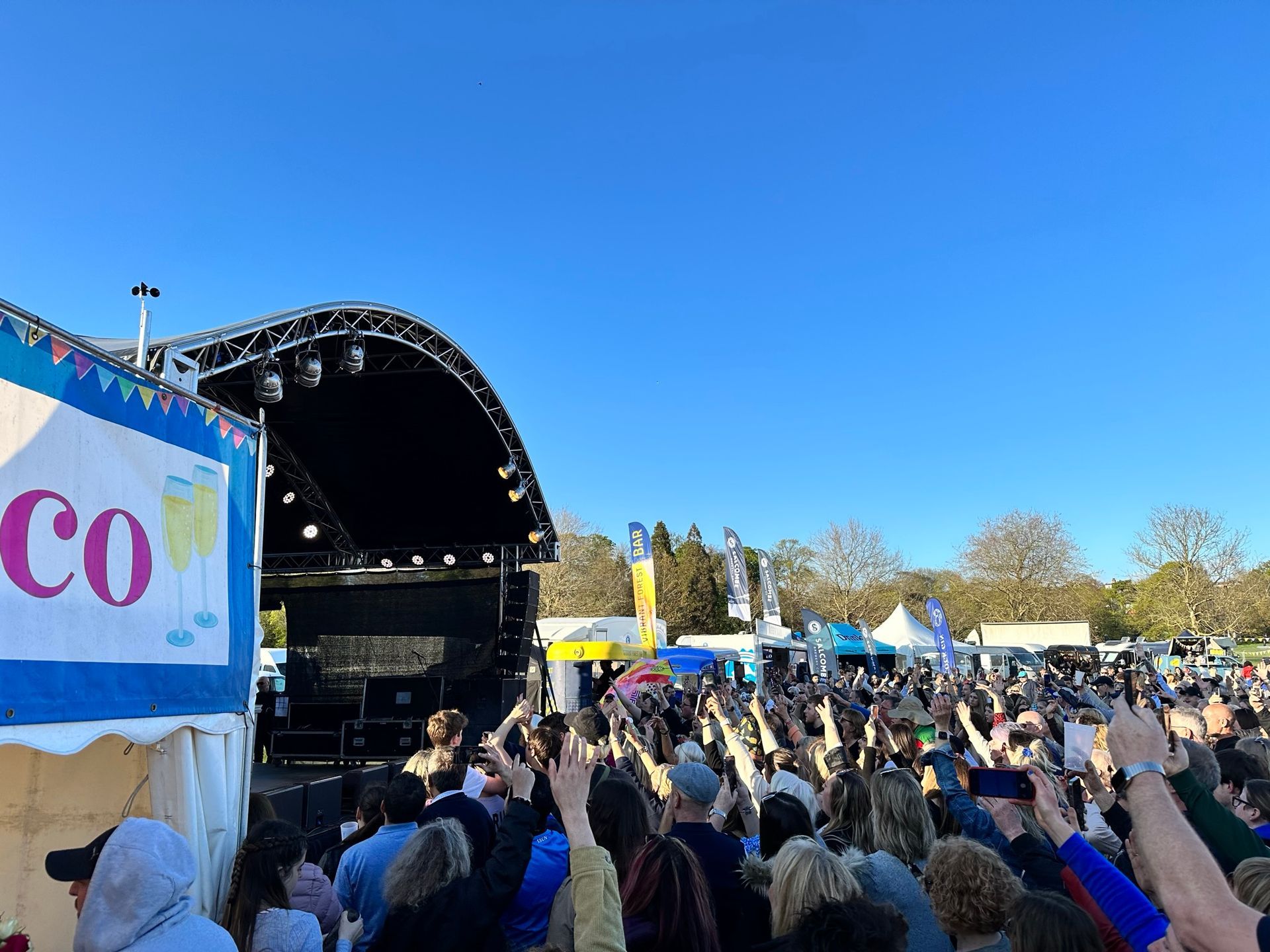 an arc truss stage below a blue sky with a large audience in front