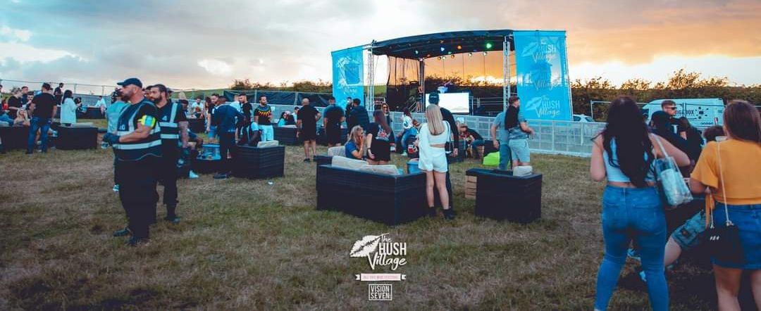 A socially distanved event in full swing, with a trailer stage set up and an audience watching in small groups. The sun is setting on the horizon and the light is fading. The stage PA wings are decorated with banners.