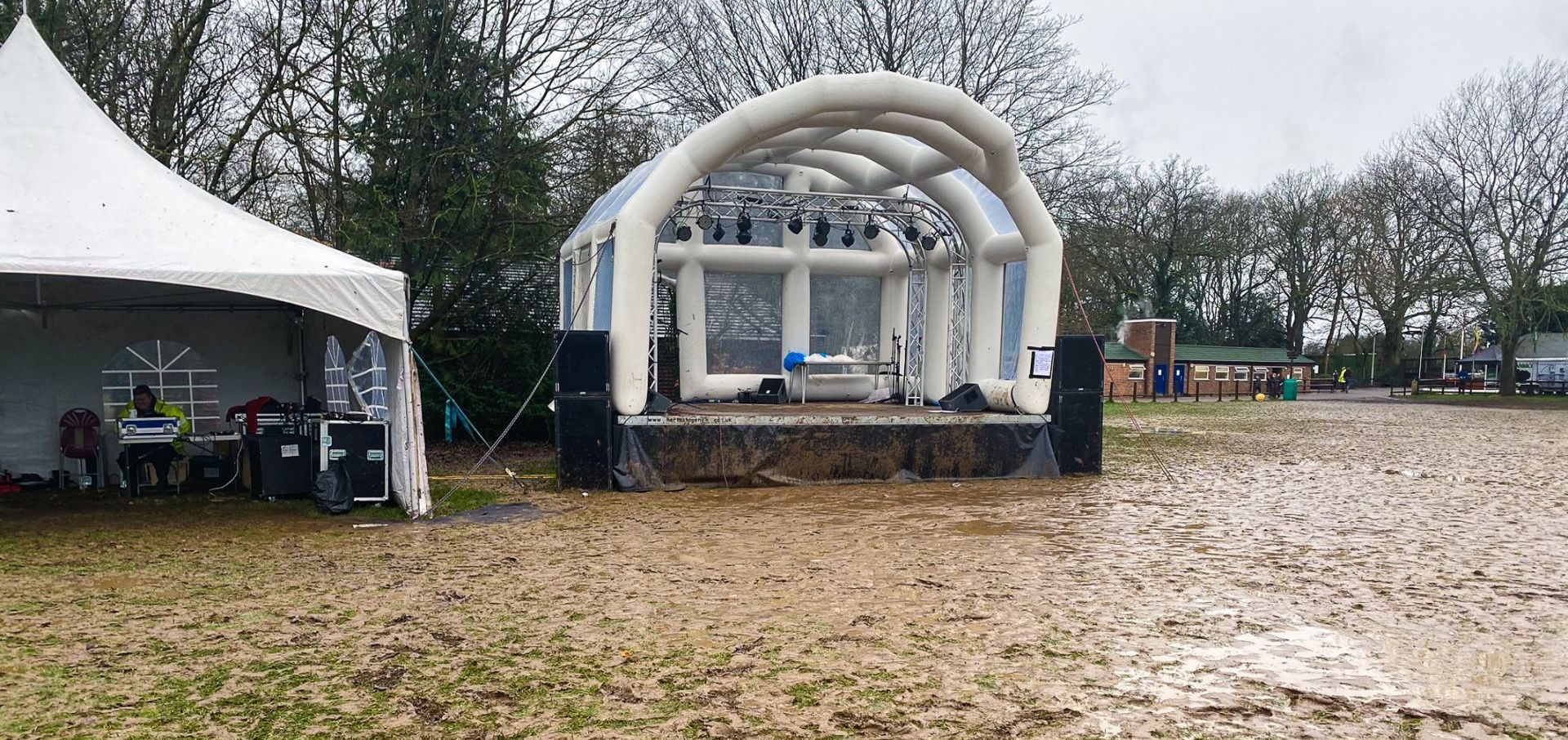 An inflatable roofed trailer stage in a very muddy and wet field, set up for an event. The black skirt at the front is covered in mud and the ground is water logged.