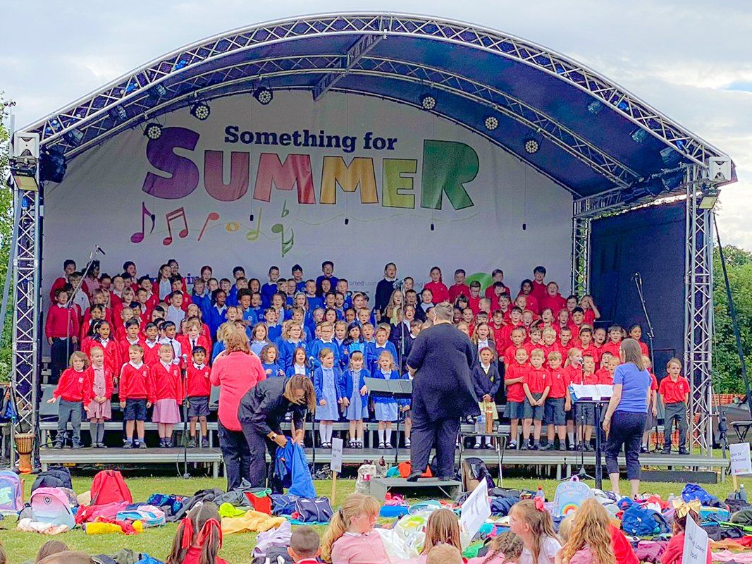 Arc truss stage with a childrens choir performing on it in front of a large audience. It's colourful with the kids dressed in red and blue and a large sign reads Something for Summer on the back wall of the stage.