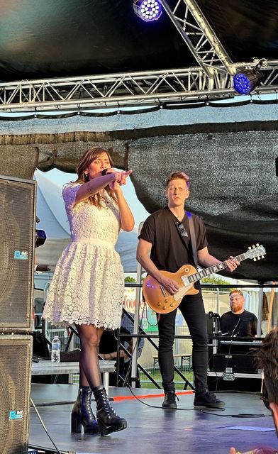 A close up shot of Natalie Imbruglia performing on an arc truss stage from Koncept Productions, with a guitarist. There are blue lights above and she wears a white dress and black books. The photo is from the foodies festival 2023