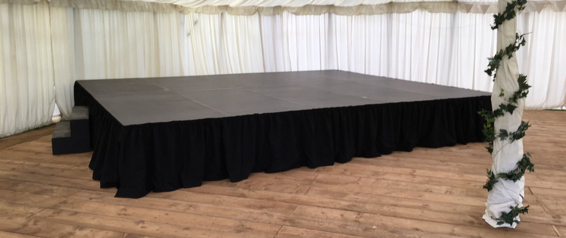 A black low level stage set up in a marquee for a wedding, with a black skirt around the edge and steps to one side. The floor is wooden and white drapes hang from the walls.
