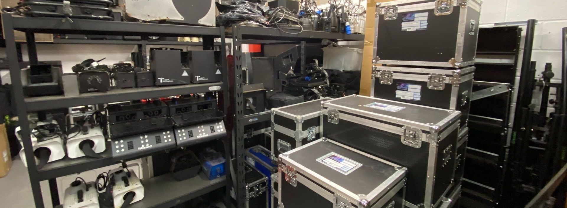 A photo of the inside of a warehouse, with equipment stacked on shelves and flight cases in the foreground.