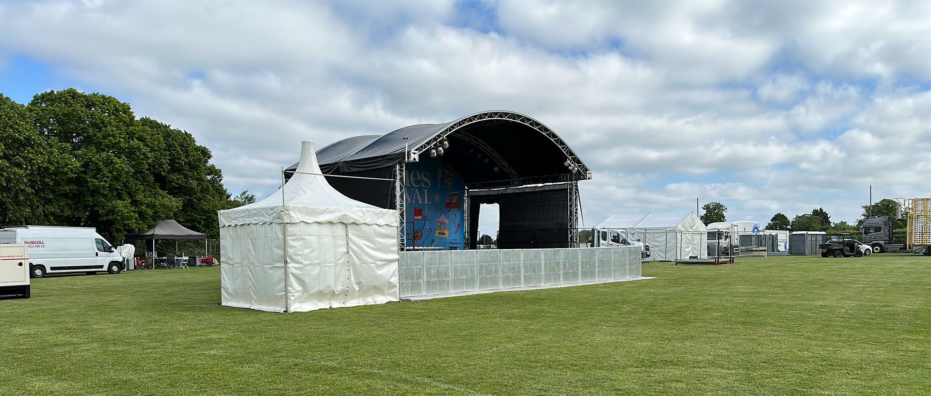 An arc truss stage in the middle of a field with a white marquee beside it