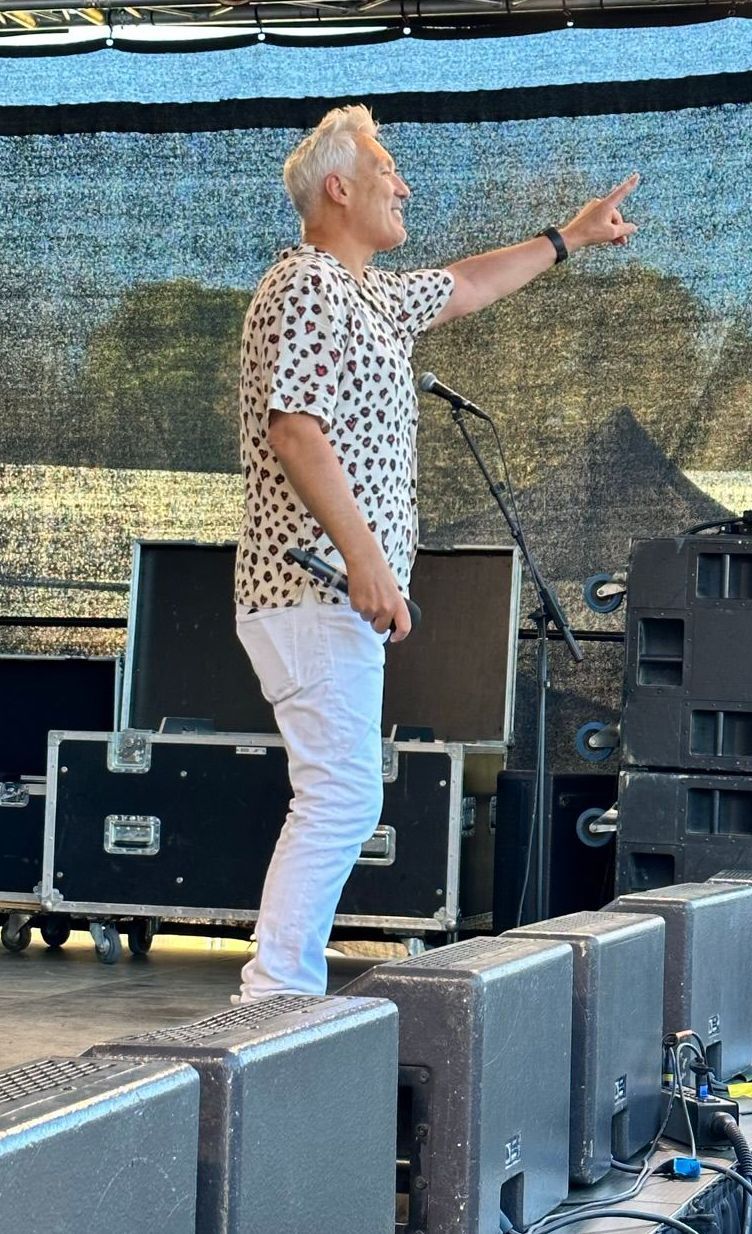 Martin Kemp interacting with the audience from an arc truss stage with production equipment in front of him on a sunny day at a festival.