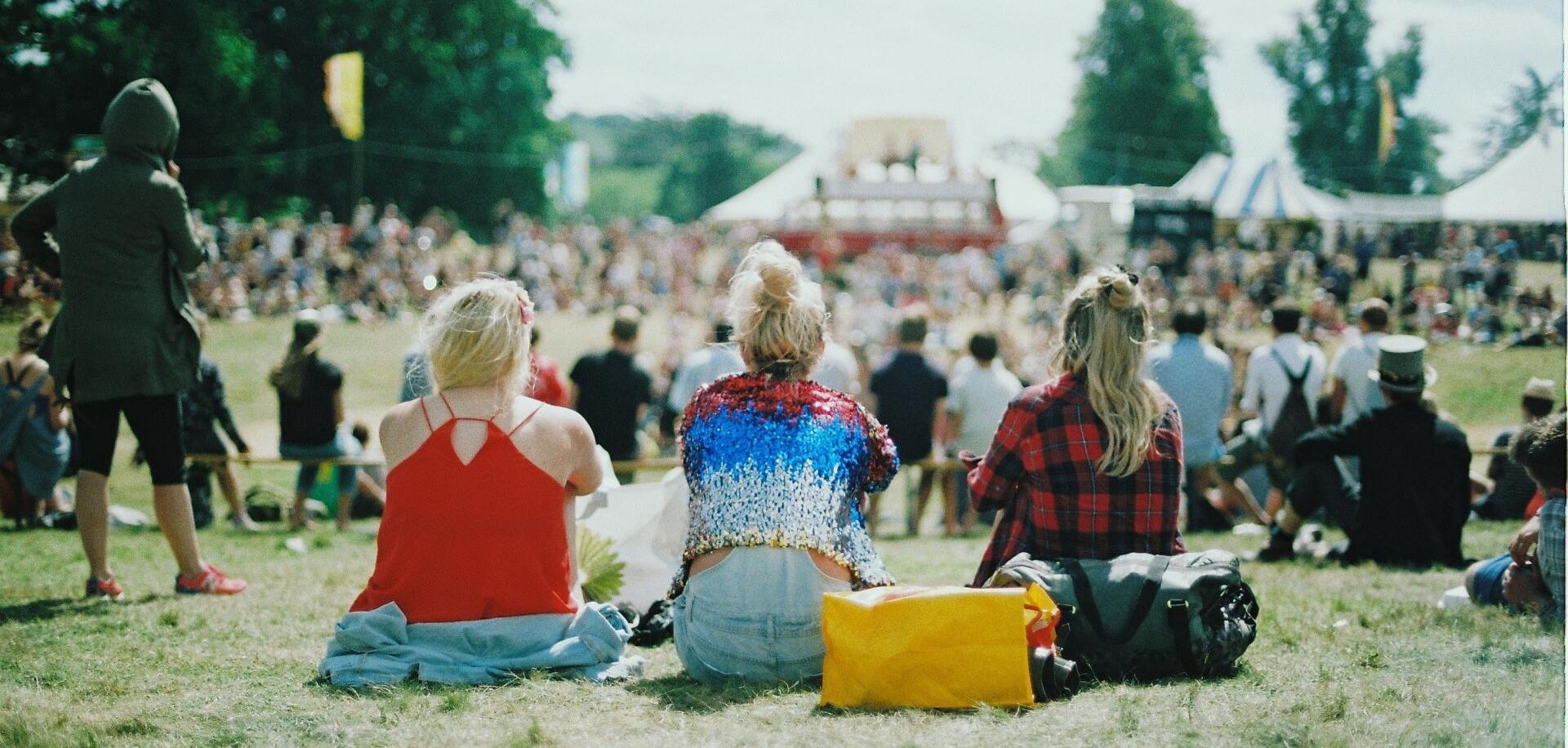 A shot from behind of three girls at a festival sat on the grass, looking over a busy festival ground that is blurred in the background.