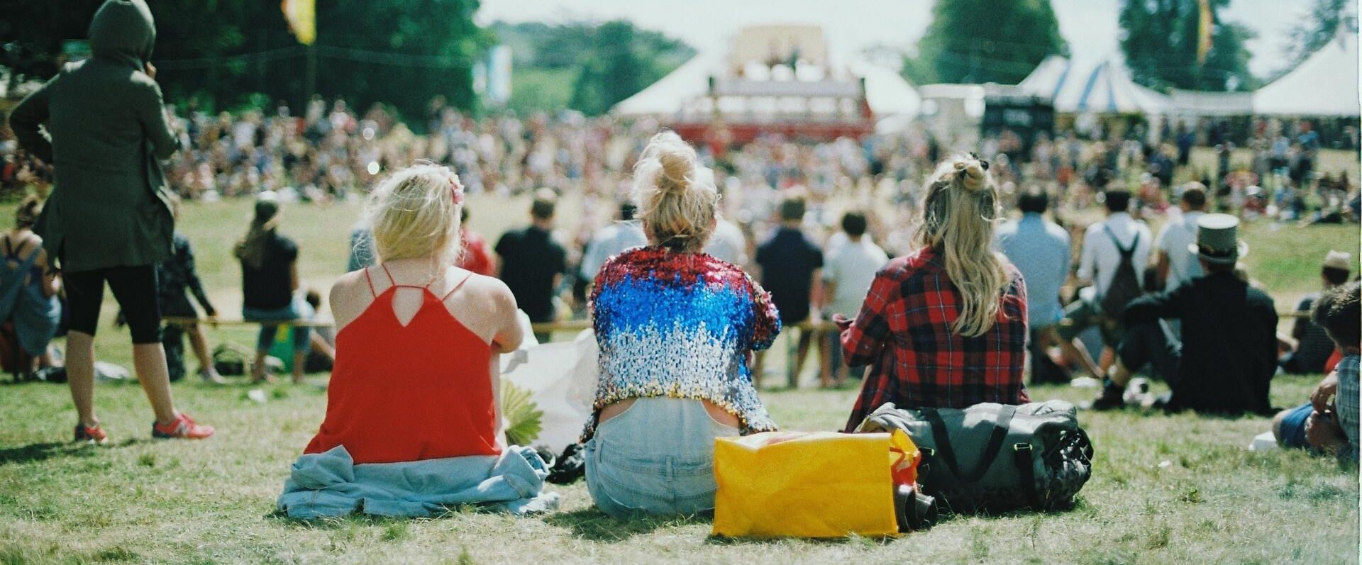 Shot of three girls backs sat on the grass with a festival in the background