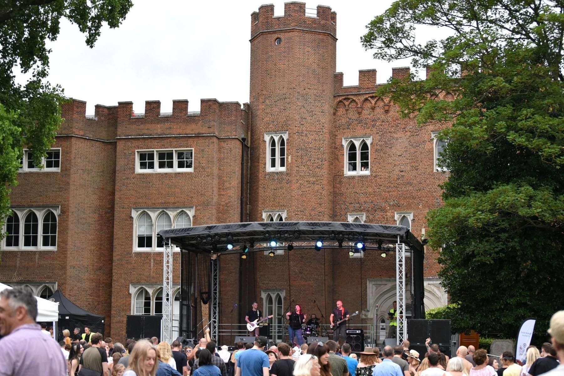 Large trailer stage set up in front of a castle, a band performing on it and a large audience in front.