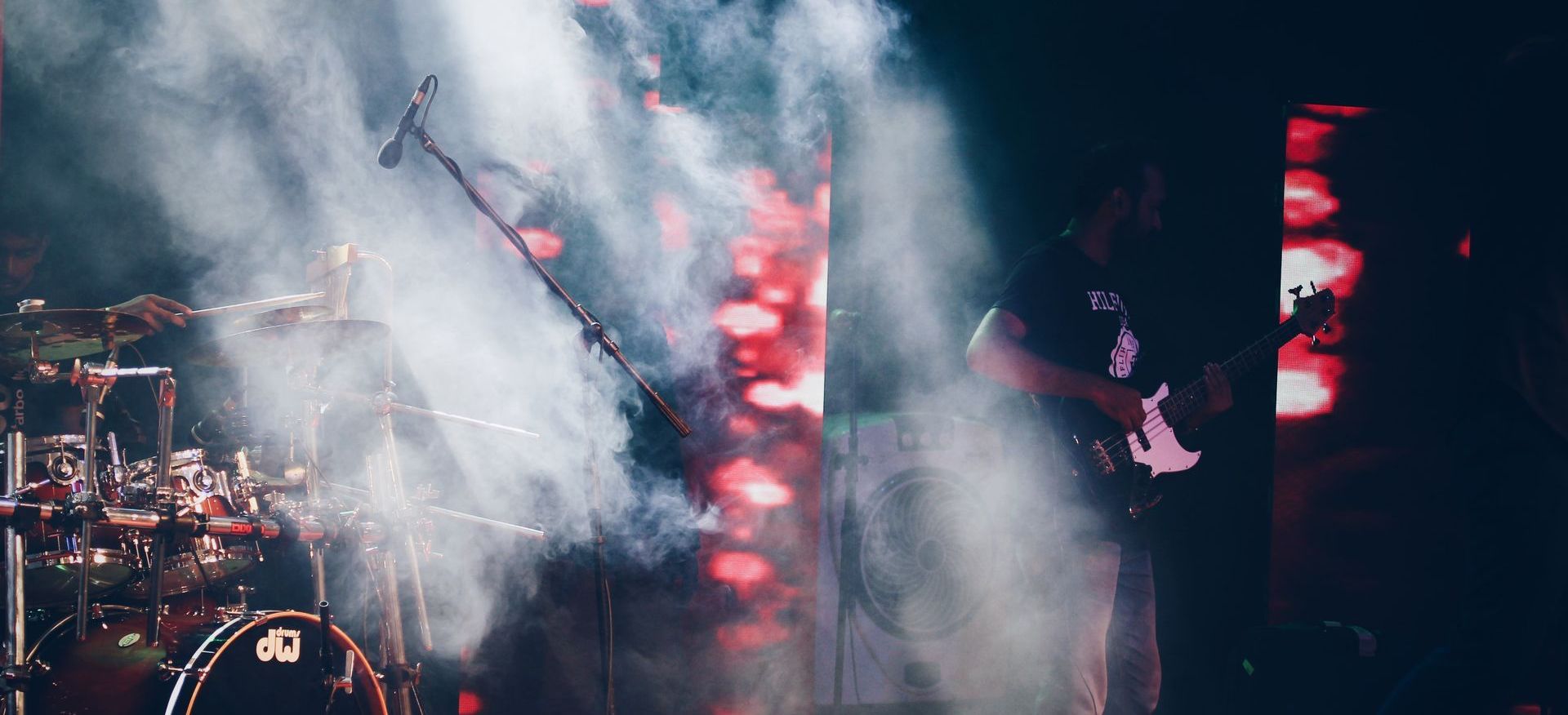 a drum set and guitarist bathed in smoke special effects and light cutting through them.