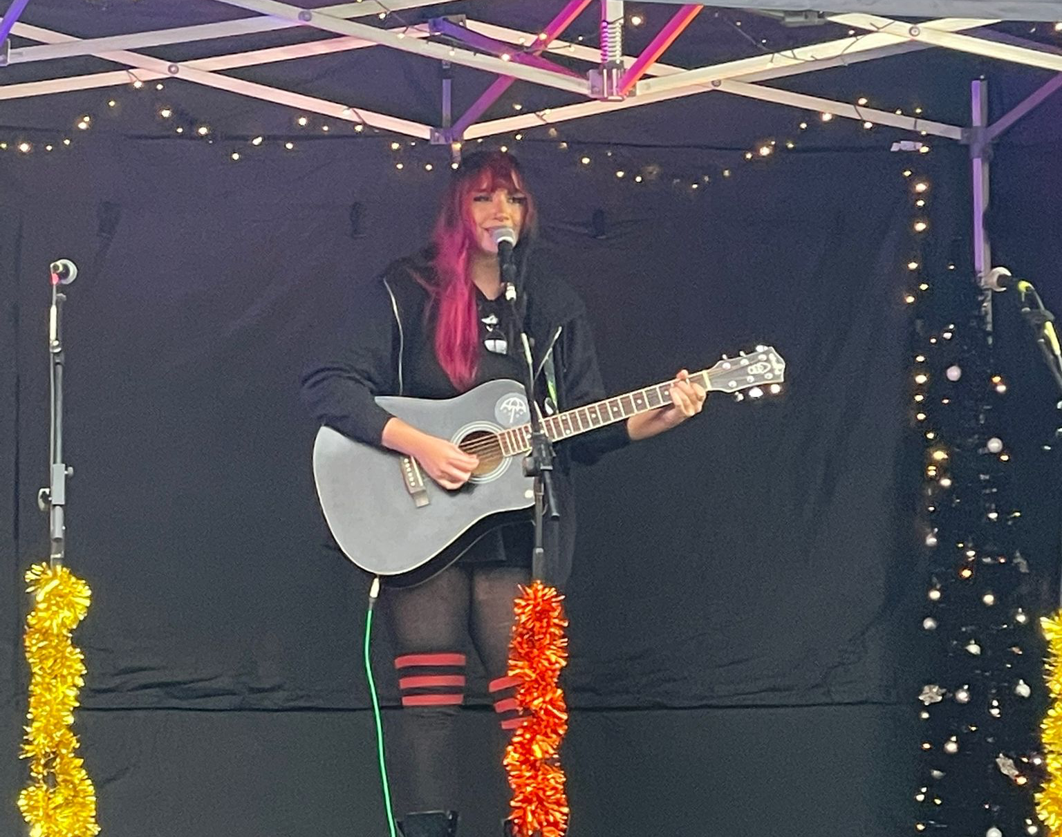 A girl with pink hair playing the guitar and singing on a modular stage at a community event