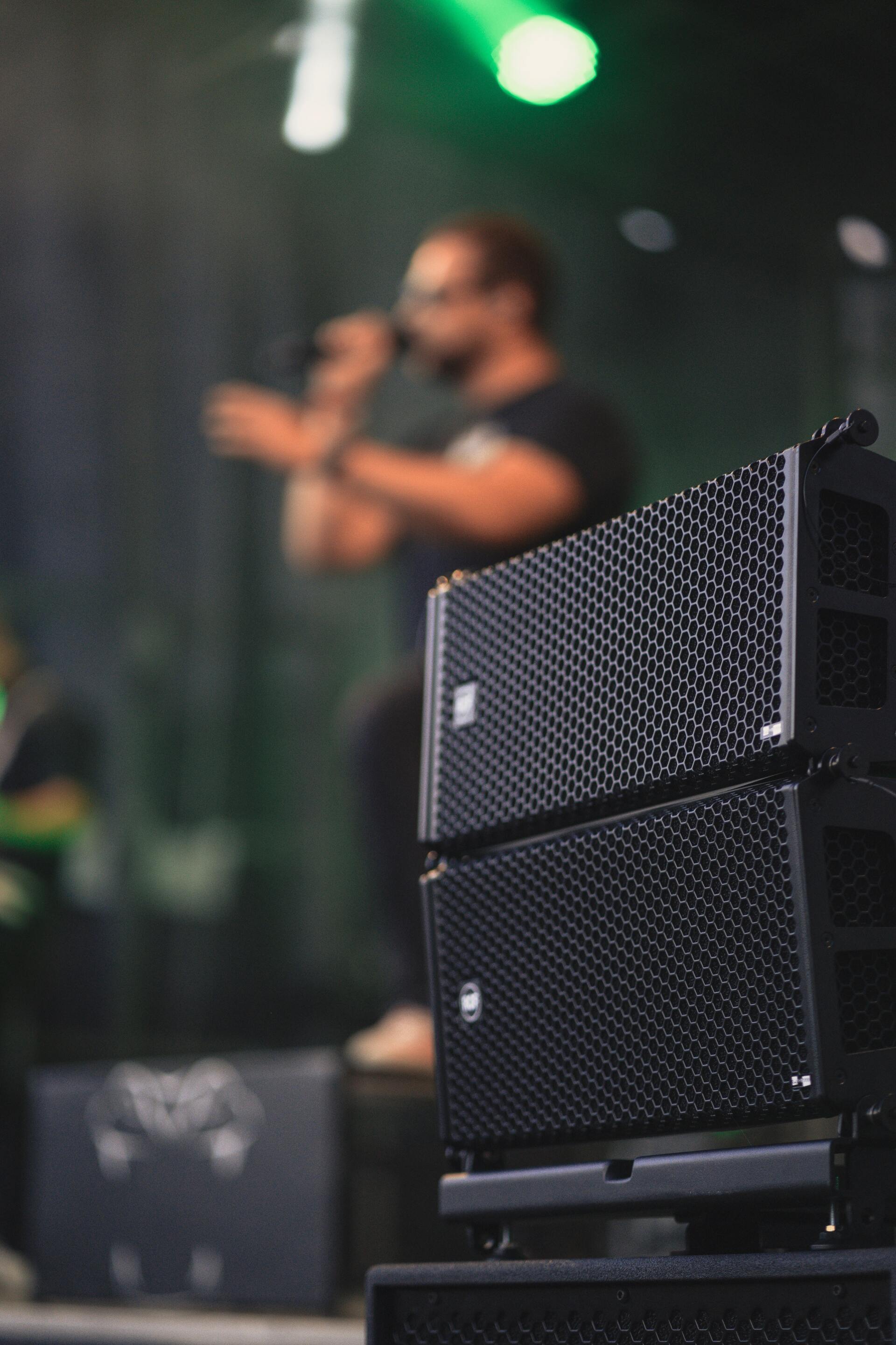 A close up of stage speakers with a singer in the background singing into a microphone.