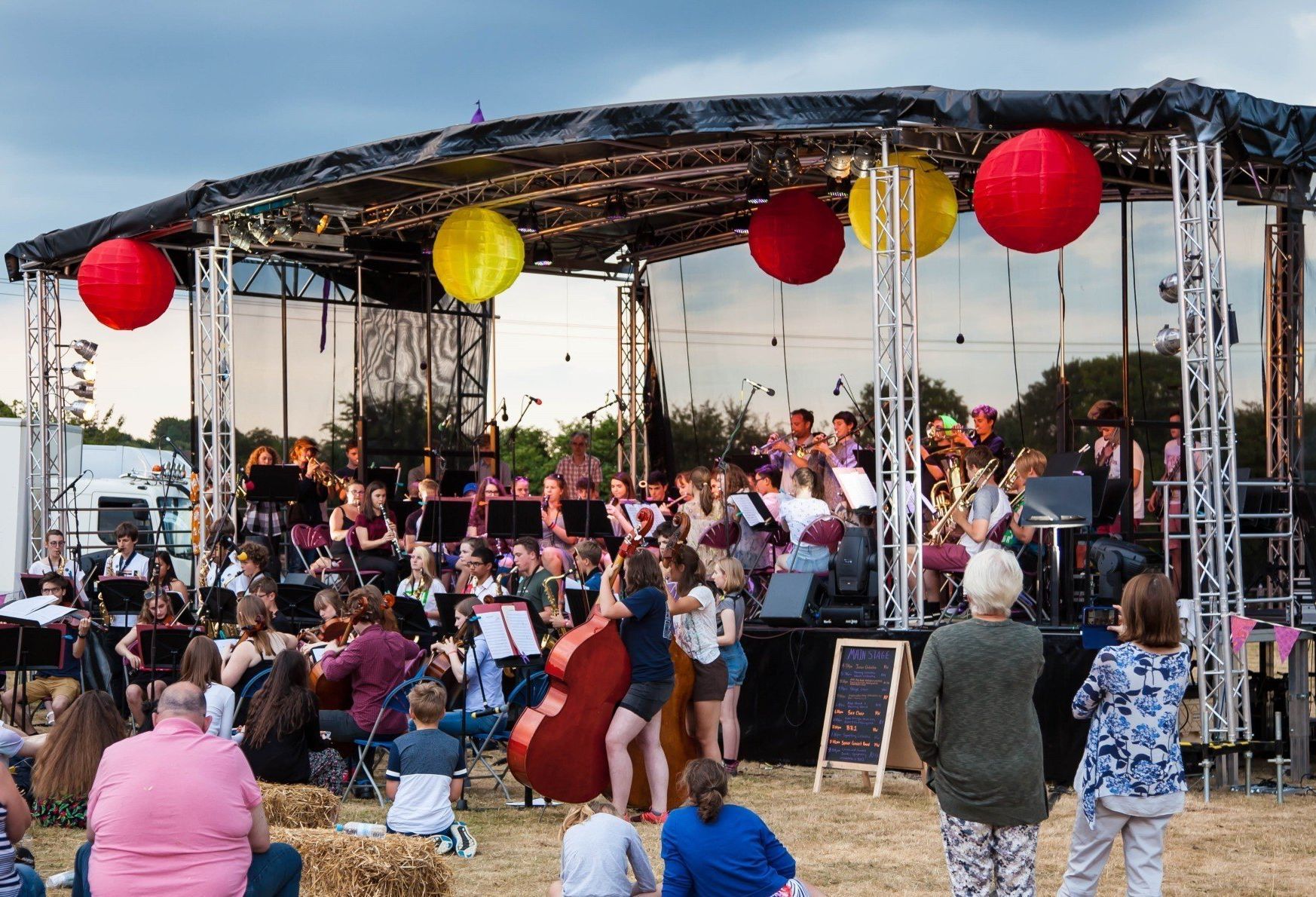 The trailer stage at a summer Gala event. An orchestra is on and around the stage where lanterns are handing and the light is fading in the sky