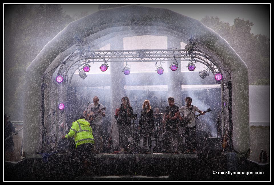 An inflatable stage with a band performing on it in the pouring rain. Purple lights hang from the truss.