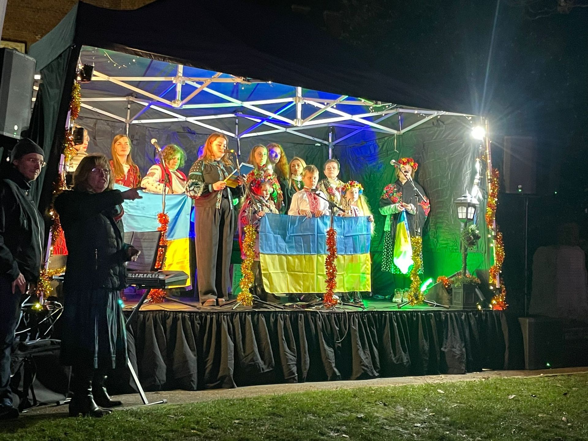 A group performing on a modular stage with festive lighting and decorations, with a black waterproof canopy whilst holding a ukrainian flag