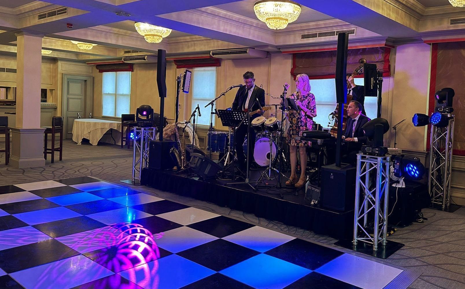 A stage set up in an inside venue for a wedding, a band playing on it, with lights and PA and a checker board dance floor.
