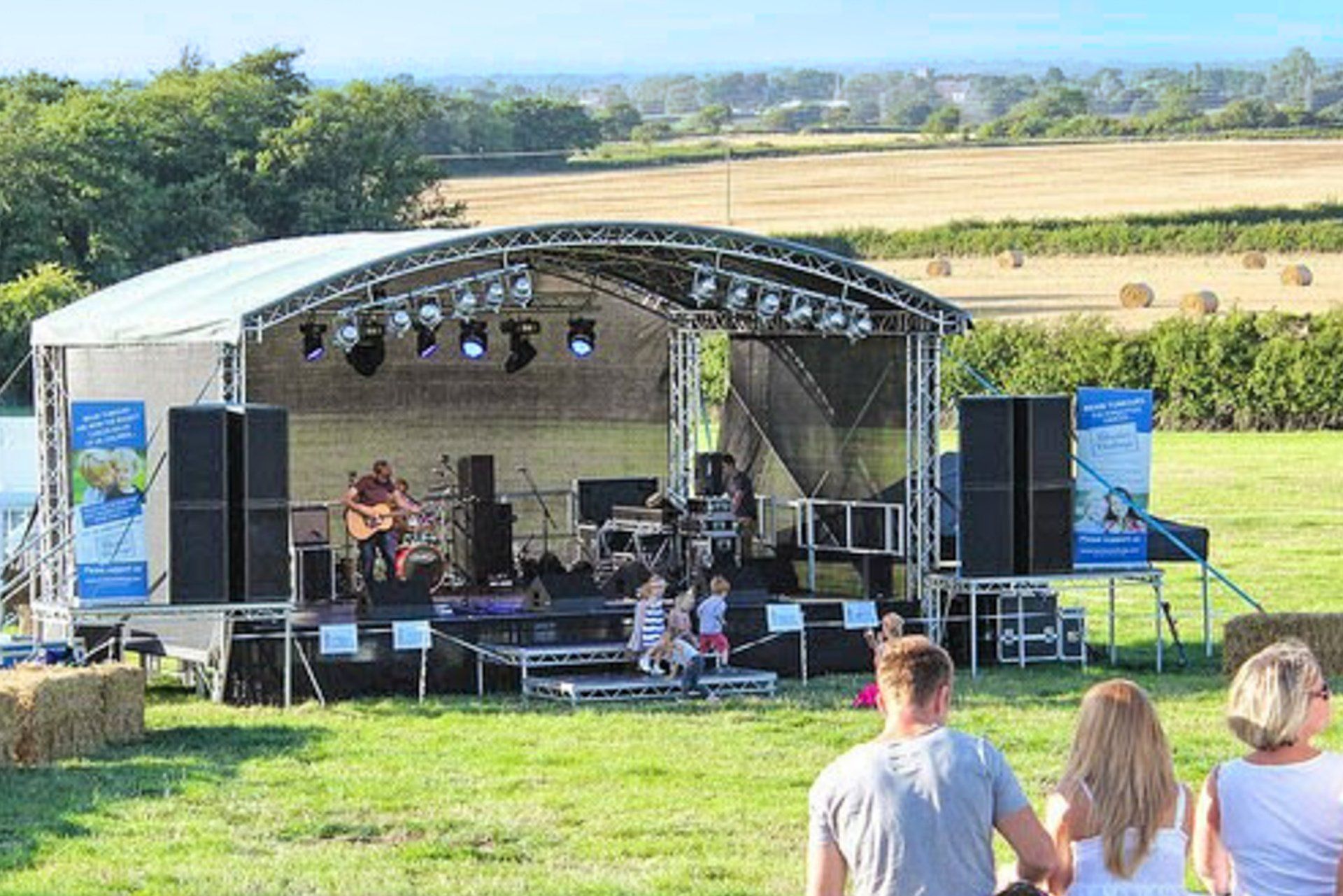 An arc truss stage in a field with performers on it and an audience watching on