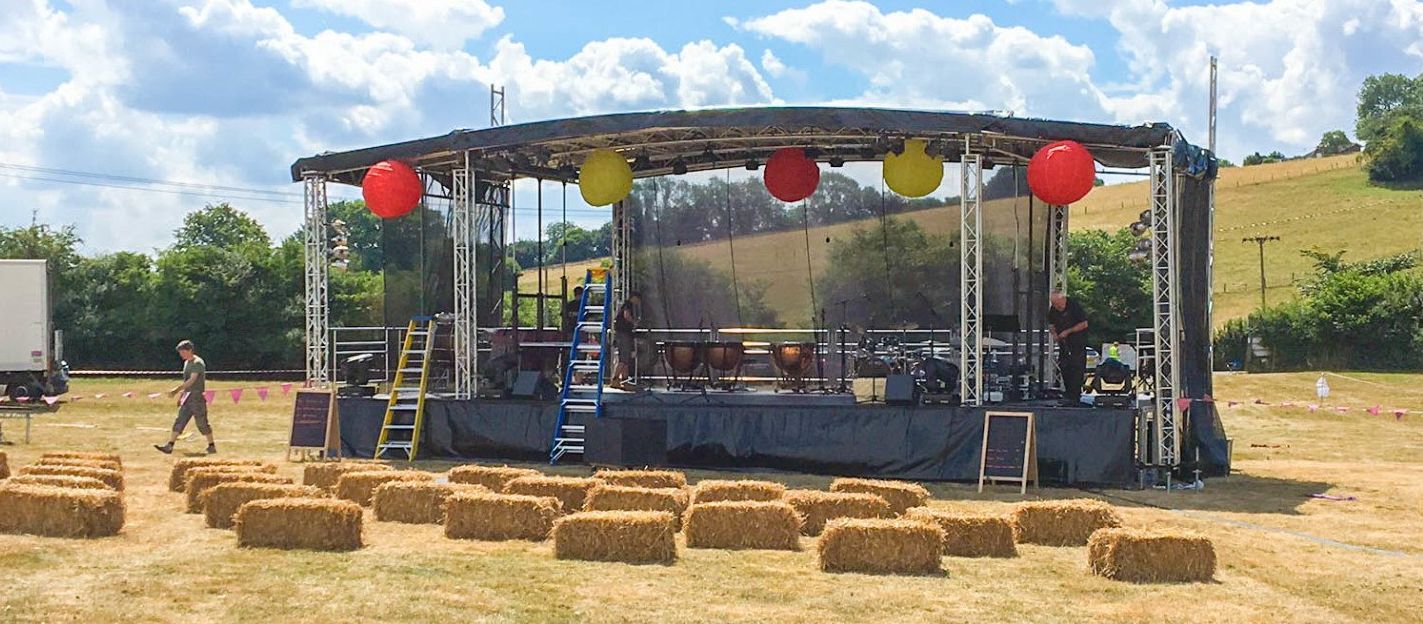 An extra large trailer stage set up in a burn out field, with red and yellow lanterns hanging from the truss and hay bails on the floor in front of it.