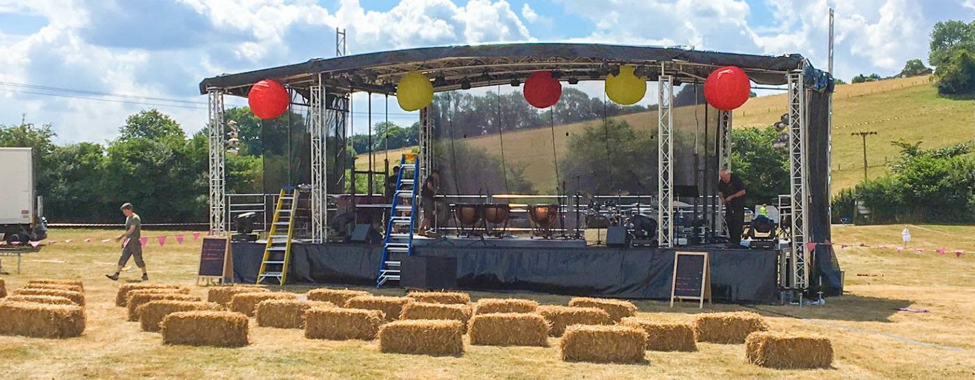 Large stage set up in a field with hay bails as seats and lanterns hanging from the trusses
