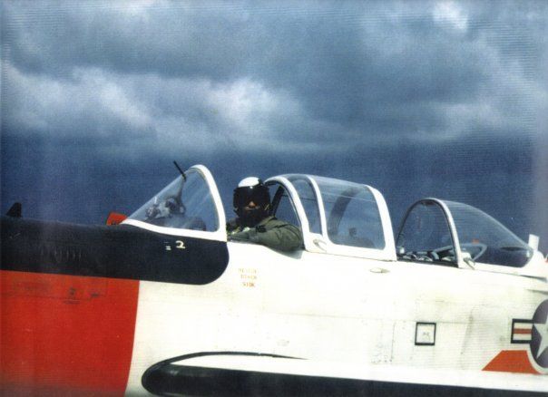 Pilot in cockpit of white and orange military training plane; dark sky background.
