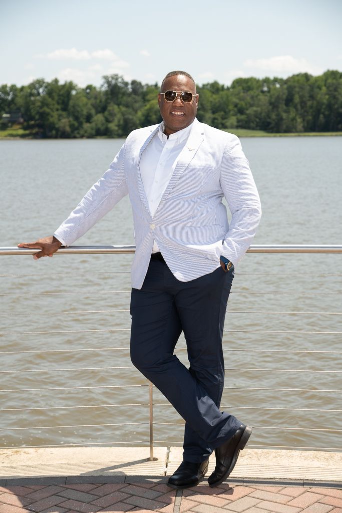 Man in a white seersucker jacket and navy pants poses by a waterfront, leaning on a railing, wearing sunglasses.