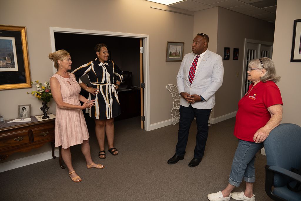 Four people in an office setting. Two women and two men are conversing, the men are standing, one wearing a blazer.