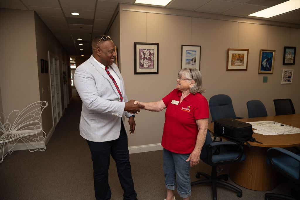 Man in white blazer and woman in red shirt shaking hands in an office hallway. Pictures hang on the wall.