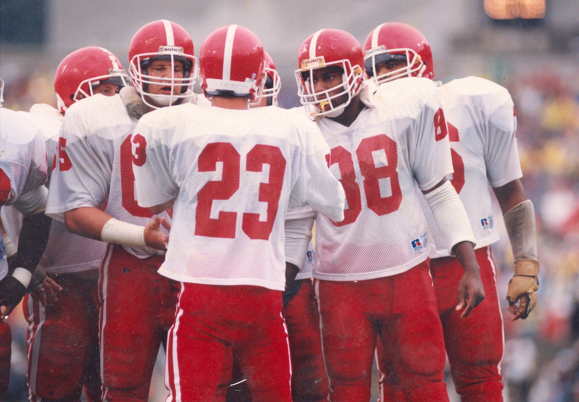 Football players in white and red uniforms huddle together on a field. Number 23 is in the center with other players around.