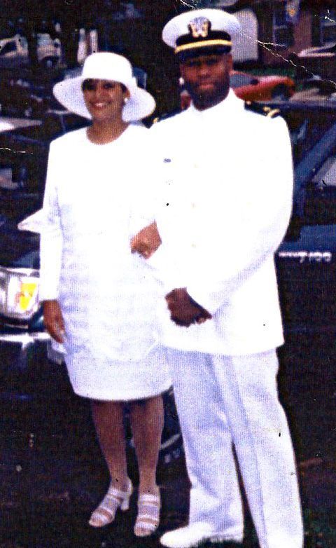 A man in a navy uniform and a woman in a white dress stand together. They appear to be smiling.