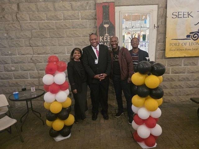Group of people posing with balloon columns at a venue with brick walls, holding a wine poster.