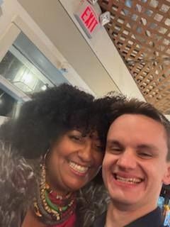 Two smiling people taking a selfie; indoors with an exit sign overhead.