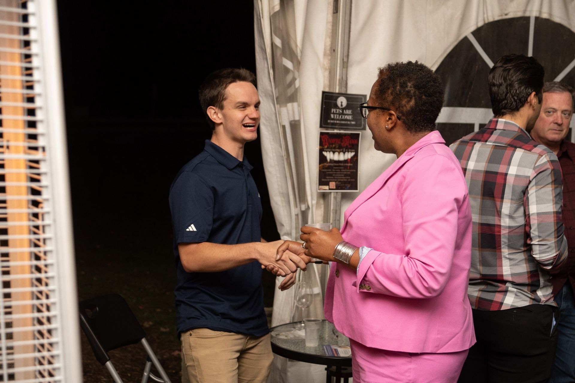 Man in blue shirt shakes hands with a woman in pink suit, other people in background.