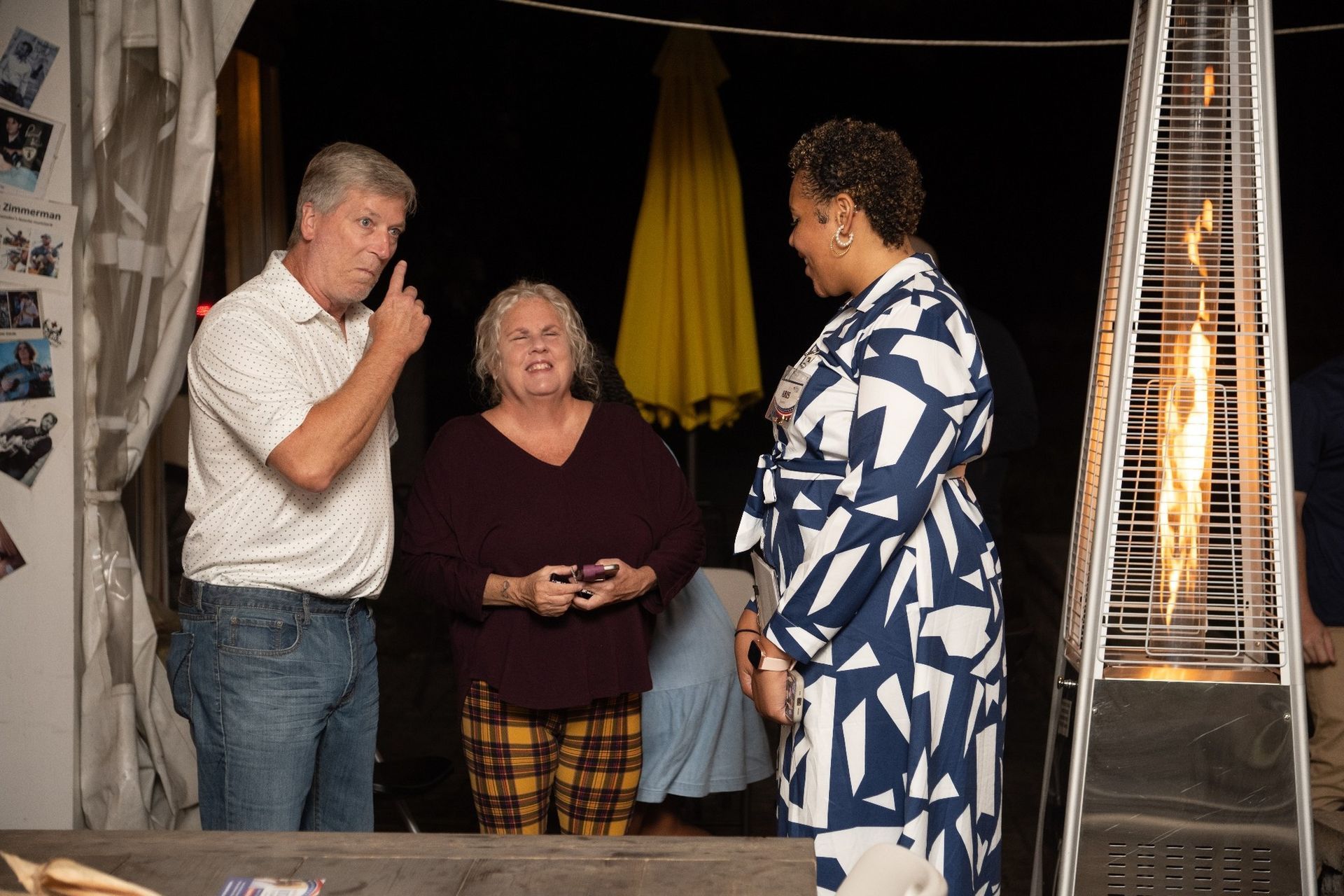 Three people talking near a patio heater. Man points; woman holds phone; woman wears patterned dress. Evening setting.
