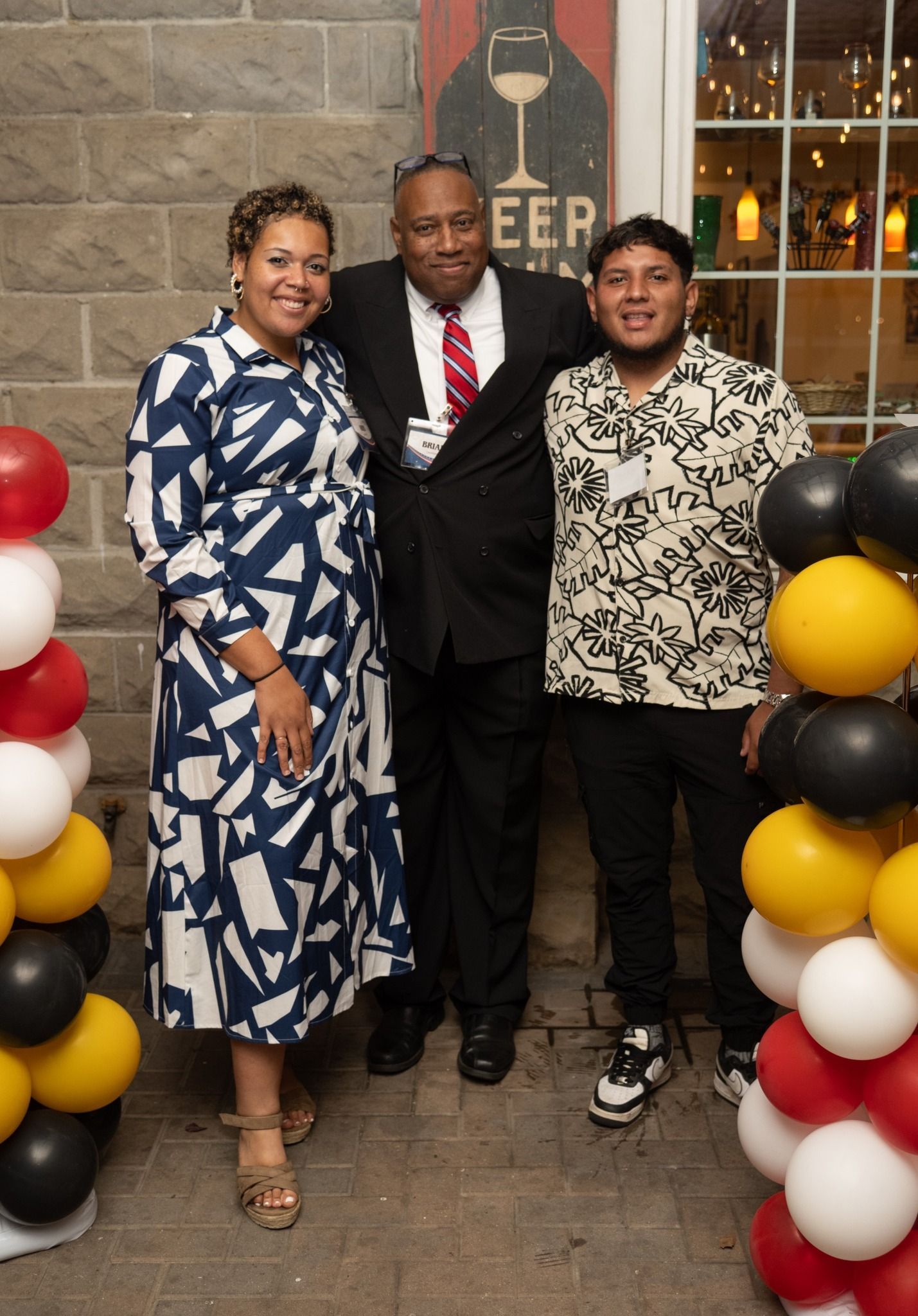 Three people pose for a photo in front of a brick wall and balloons.