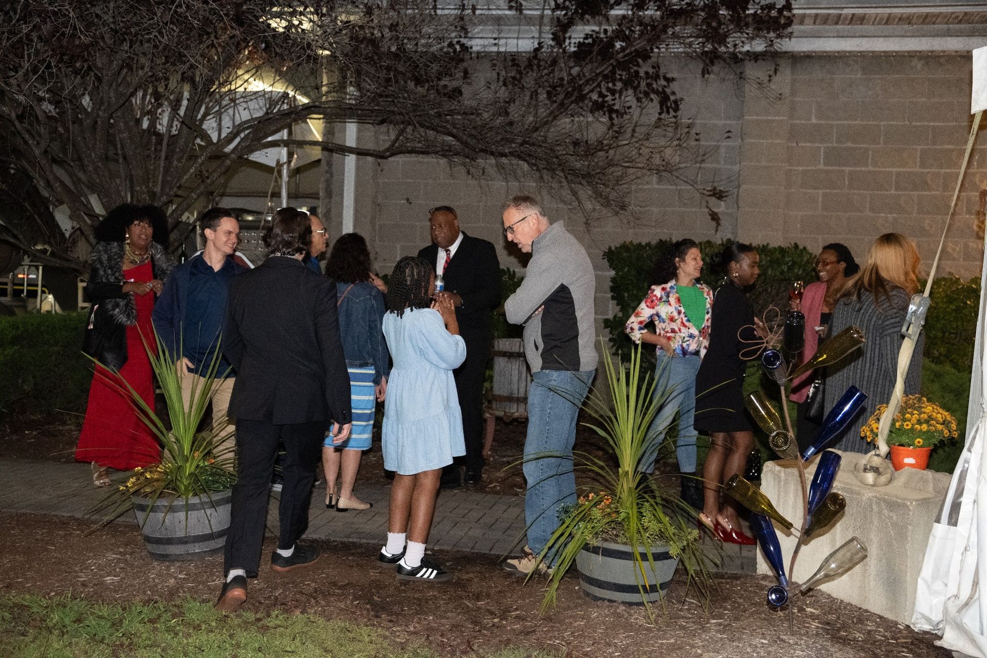 Group of people gathered outdoors, some talking, near plants and a building.
