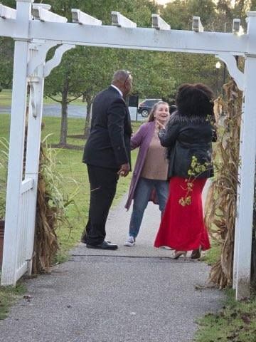 People interacting under a white arbor. One in a suit, two women, one in red skirt, one with curly hair. Park setting.