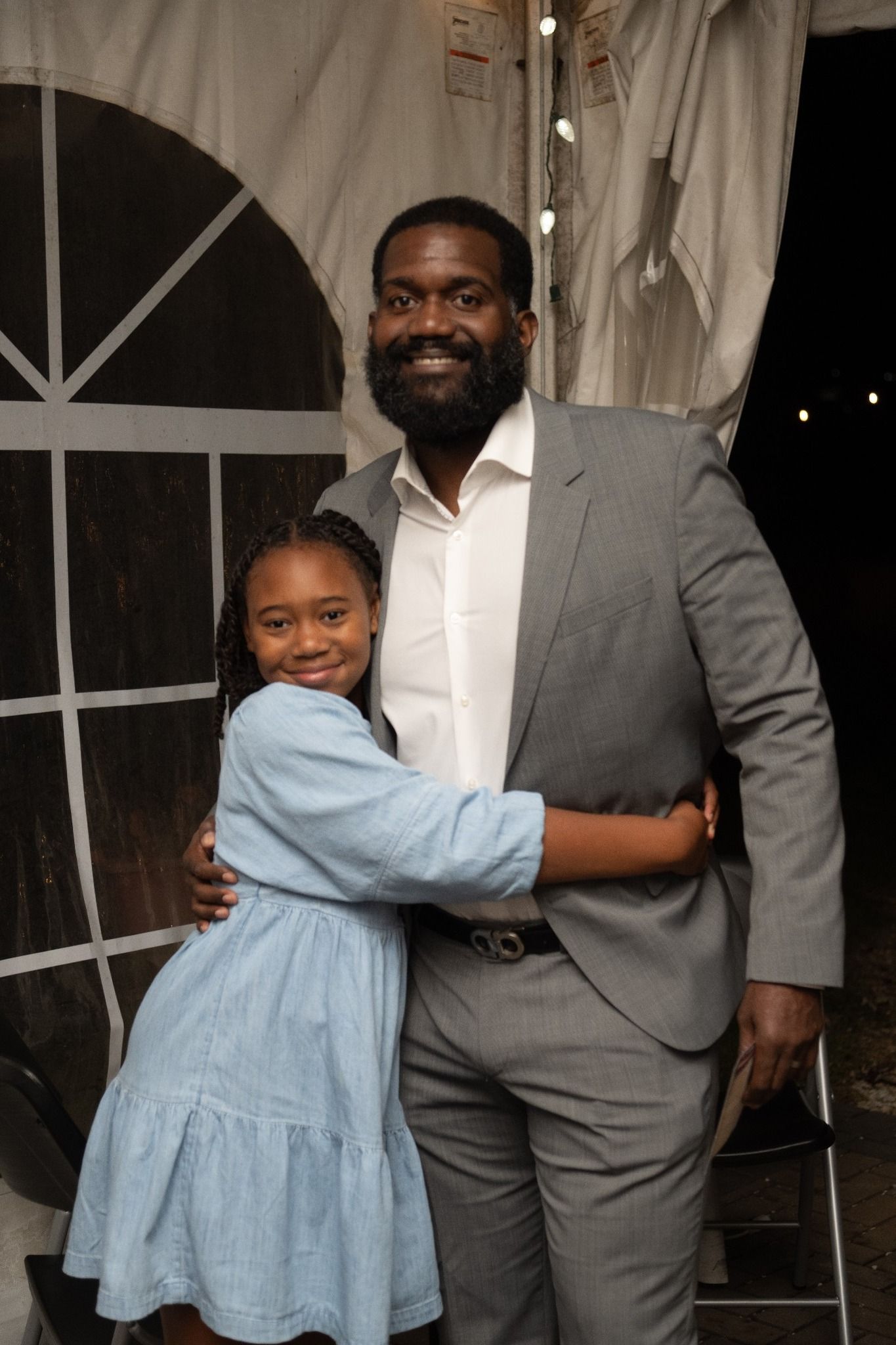 Man in gray suit and young person in blue dress embrace, smiling. Tent in background.