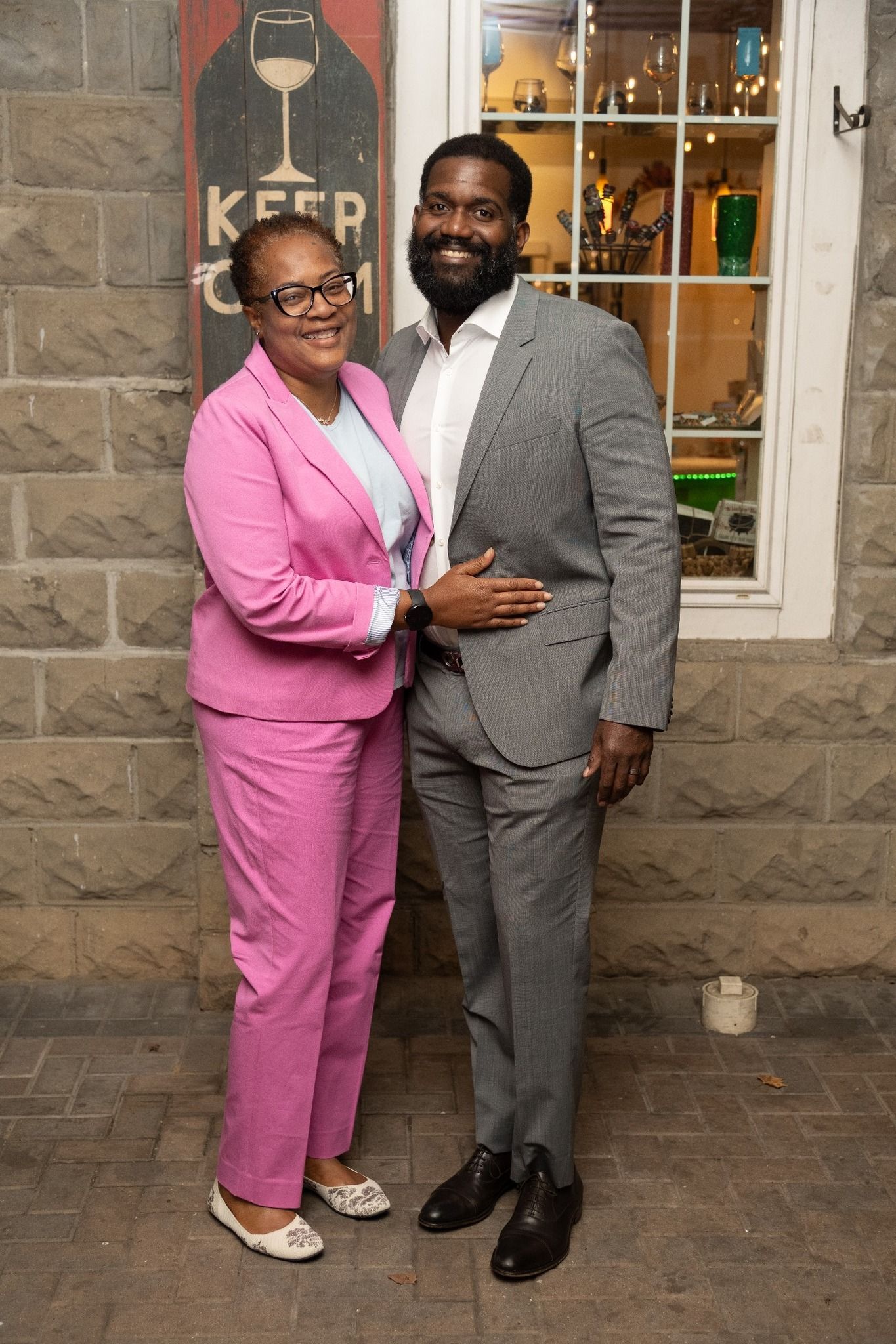 Woman in pink suit and man in gray suit standing in front of a brick wall, smiling.